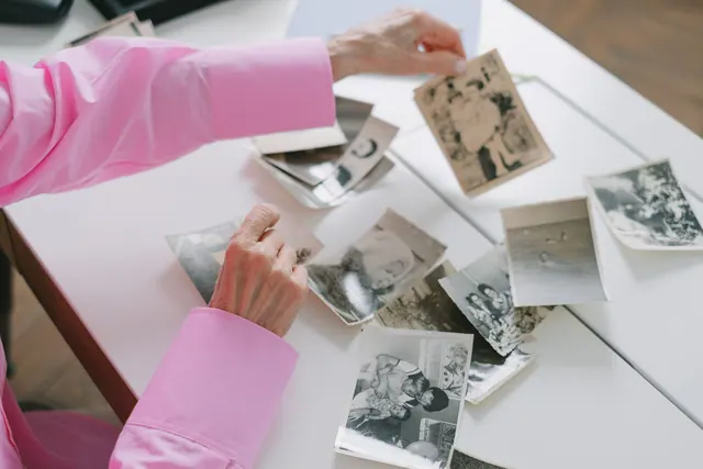 An elderly woman in a pink shirt sorting through old black and white family photographs, evoking nostalgia. People by heritage list: a researched guide